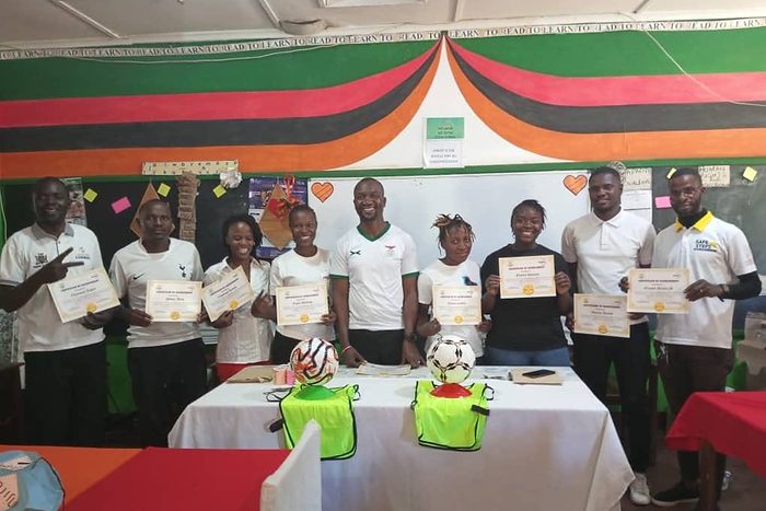 Coaches and staff stand in a classroom holding certificates, with footballs and training bibs on the table in front of them.