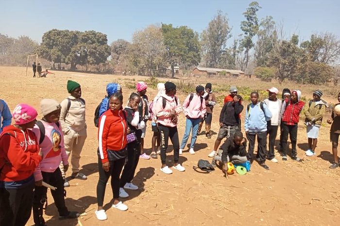 A large group of girls gathers with bags and kit on a bare, dusty pitch at the edge of the trees, ready for a session.