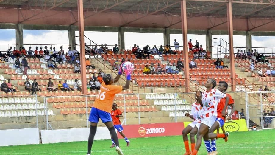 A Chipata Girls FC goalkeeper rises to claim the ball above a crowd of players, in front of a stand of watching supporters.