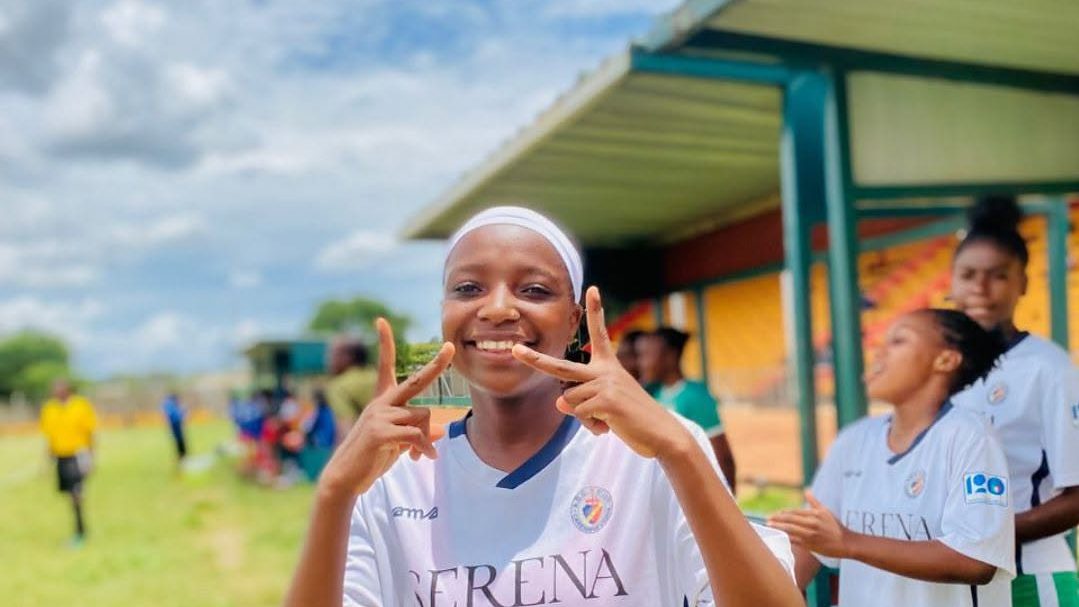 A Chipata Girls FC player in white kit smiles broadly on the touchline on a match day, teammates warming up behind her.