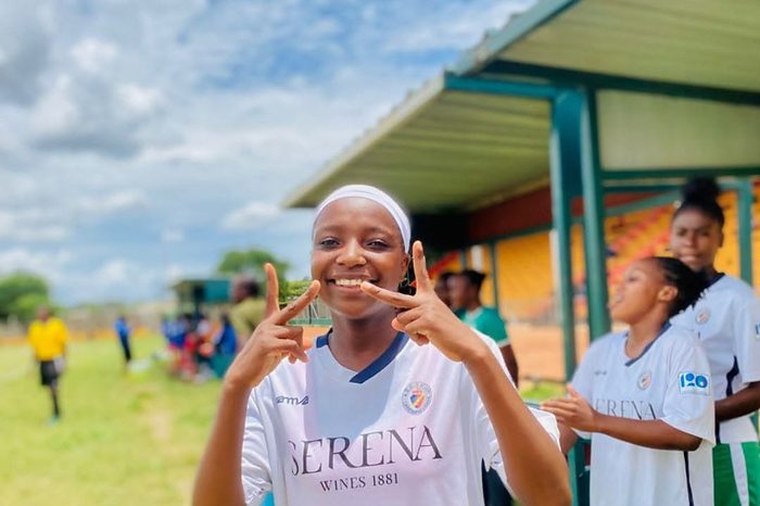 A Chipata Girls FC player in white kit smiles broadly on the touchline on a match day, teammates warming up behind her.
