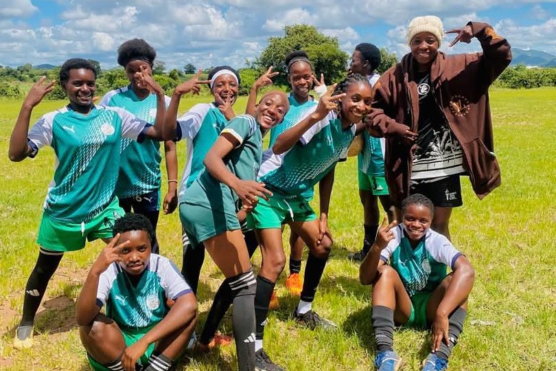 The squad grouped together on a grass pitch under a wide sky, laughing and posing for the camera after training.
