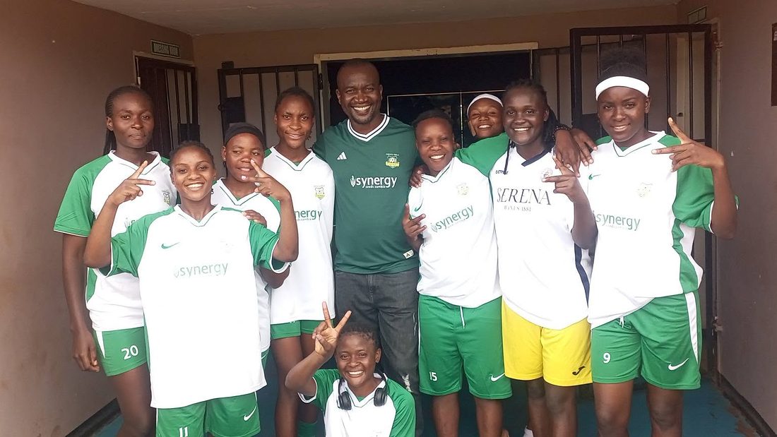 Players in club kit crowd around their coach in a doorway at the ground, arms around each other, everyone grinning.
