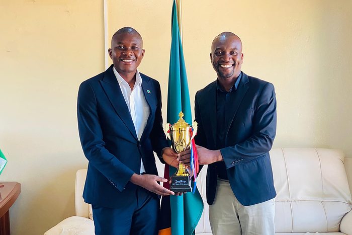 Chipangula Dube accepts a ZedPro Division One Team of the Season trophy, both men holding it between them beside a Zambian flag.